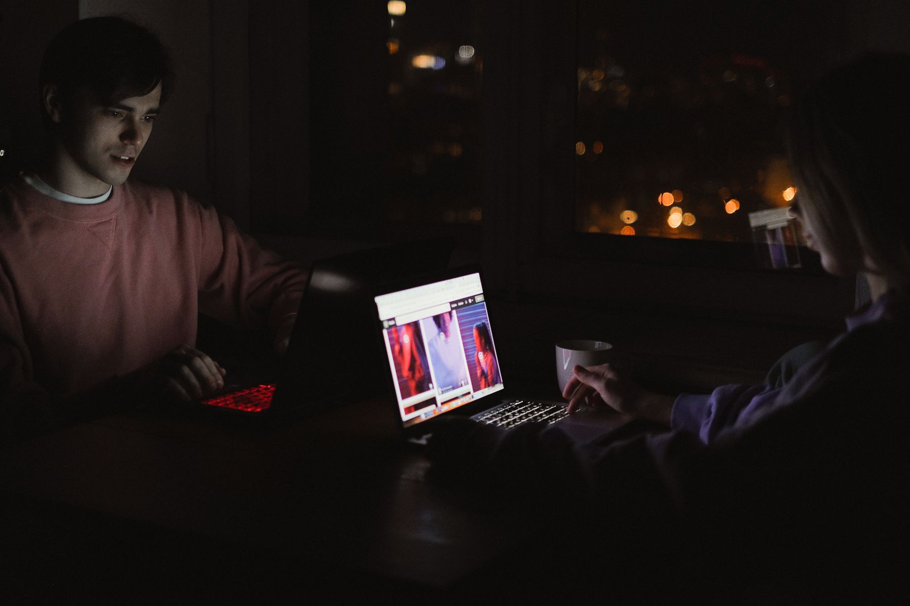 Two people working on laptops side by side at night with city lights behind them