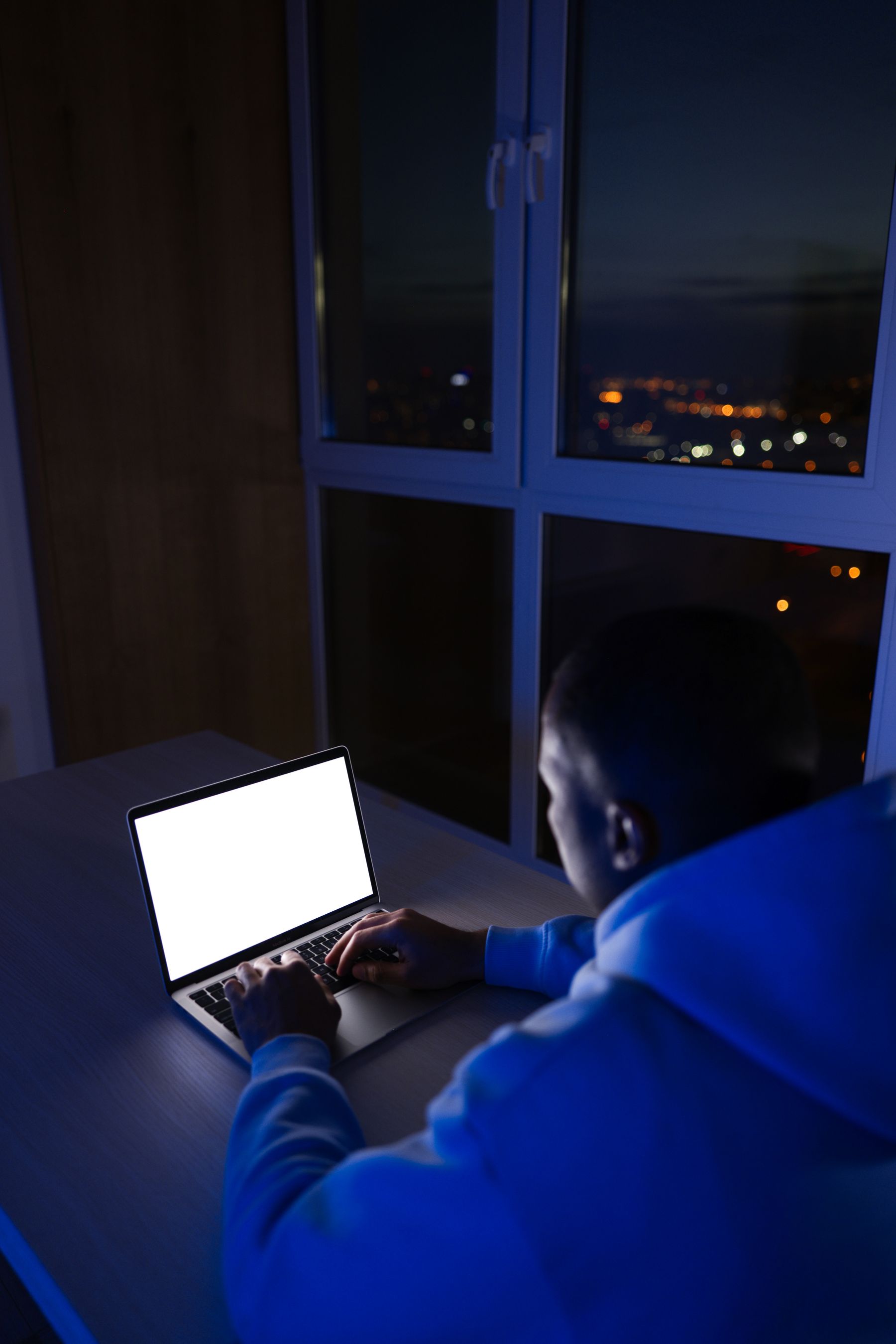 A person typing on a laptop at night beside a window over the city