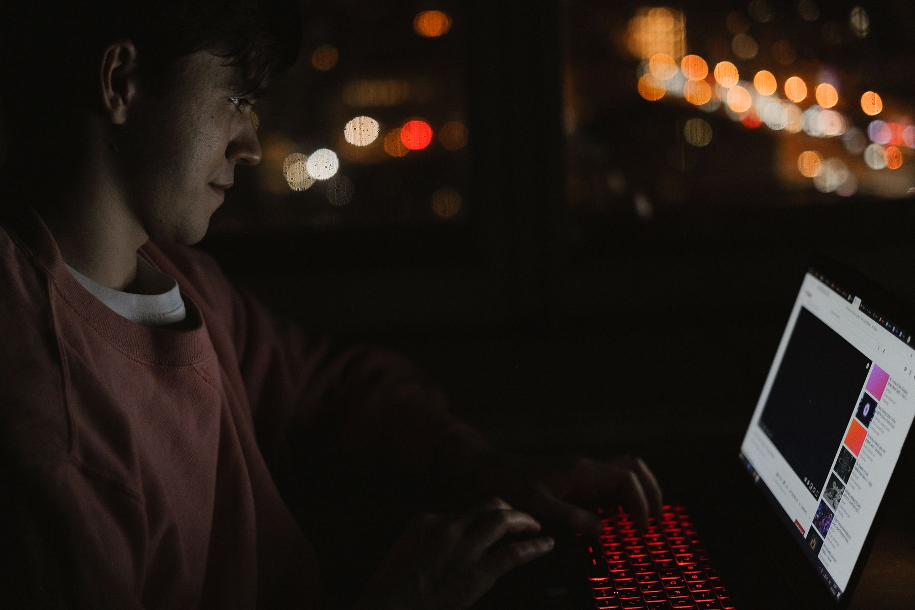 A young man working at a laptop at night with city lights blurred behind him