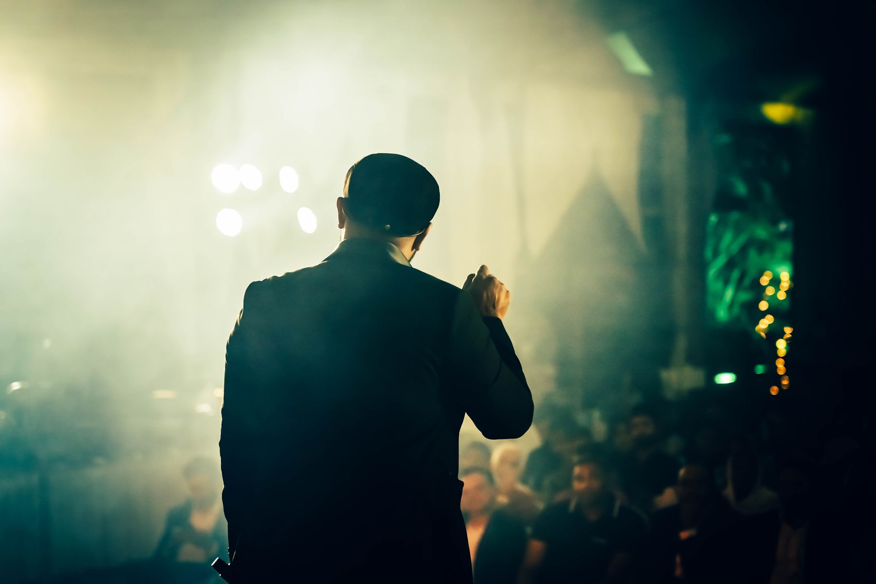 A speaker seen from behind, addressing an audience through stage haze